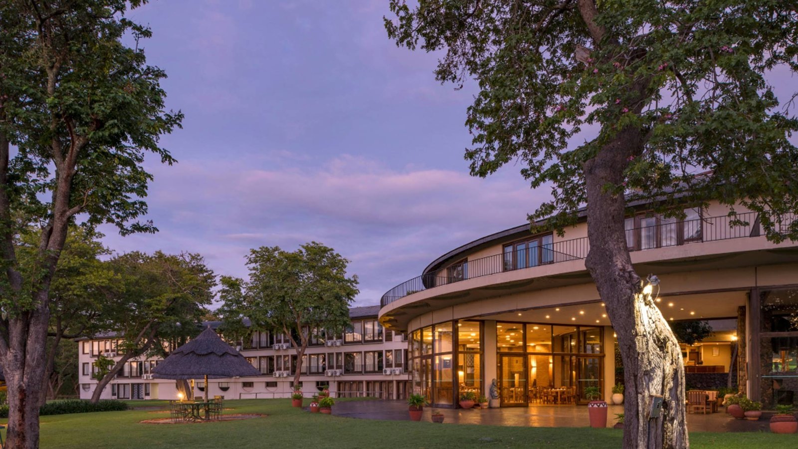 Hwange Safari Lodge's curved main building lights up at dusk beyond the lawn