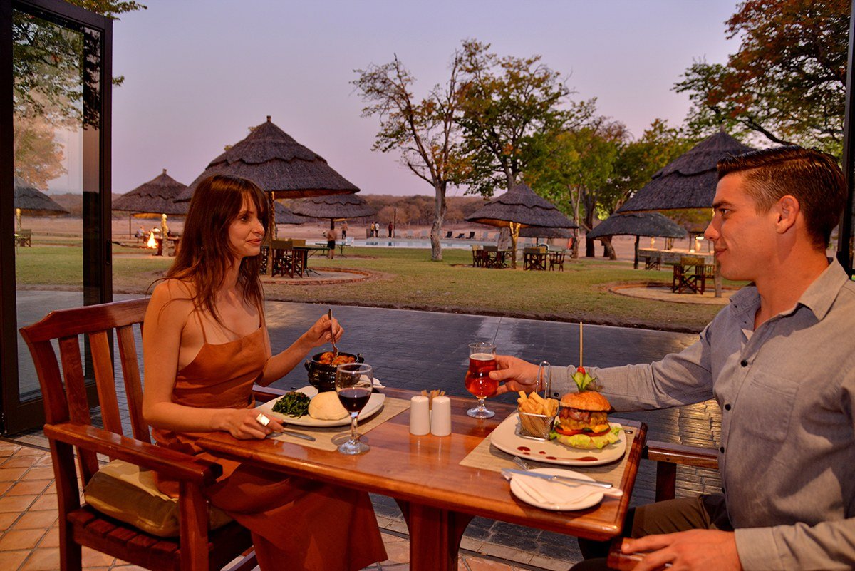 A couple dines on the terrace at Hwange Safari Lodge with thatched shelters behind them