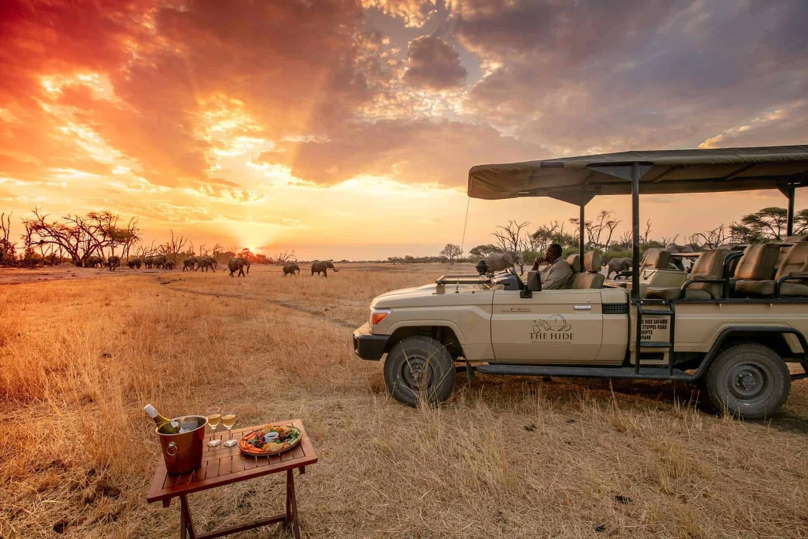 A game vehicle stops for sundowners at The Hide Safari Camp as elephants cross at sunset